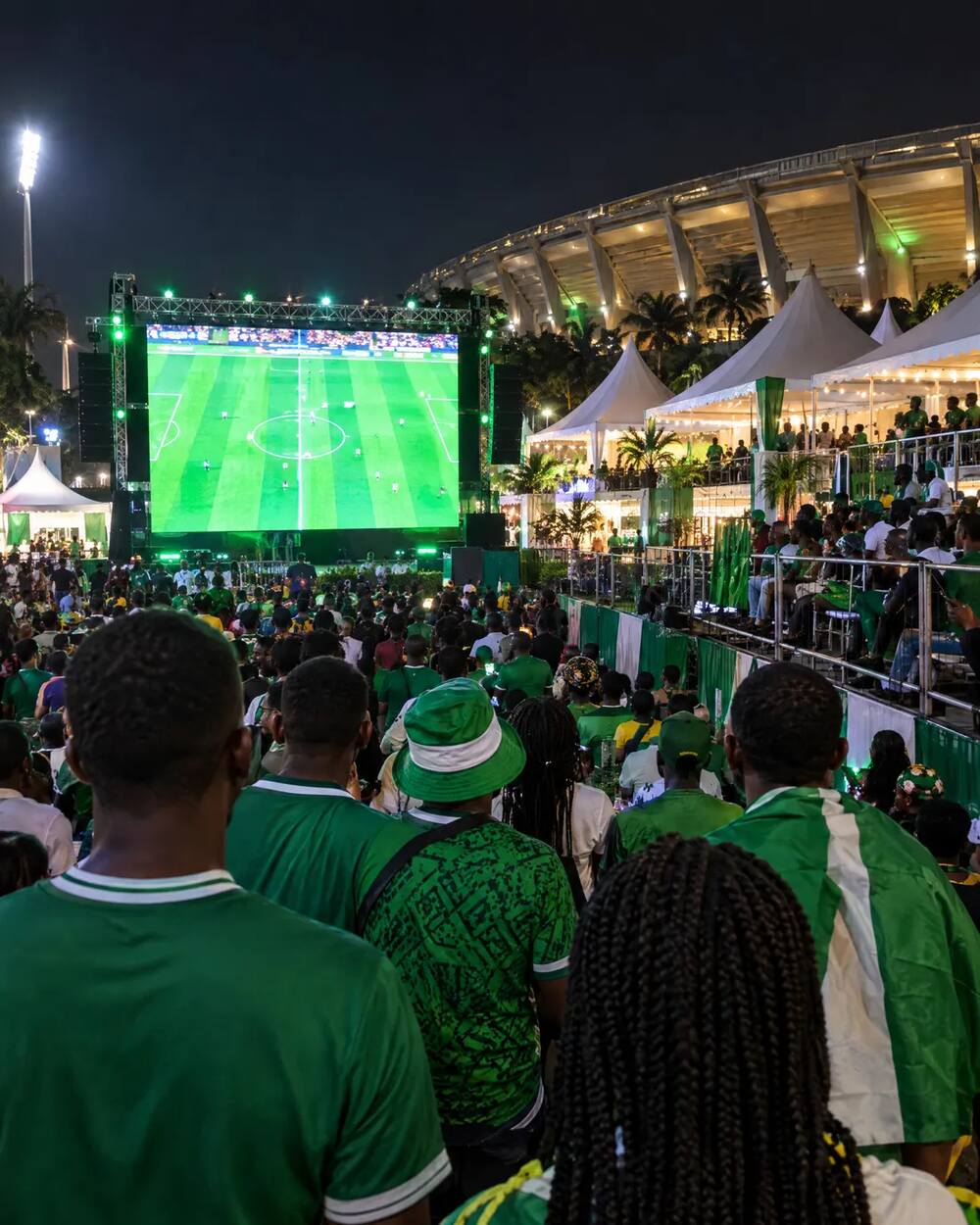 Fan zone de nuit devant un stade, écran géant et supporters en vert